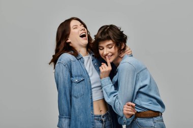 Kot elbiseli iki şık genç kadın, gri bir arkaplanda birbirlerine gülüp eğleniyorlar..