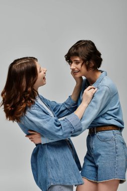 Kot elbiseli iki genç kadın, gri bir arkaplanda poz vererek, neşeli bir yakınlık ve moda anını yakalıyorlar..