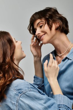 Kot gömlekli iki genç kadın gri bir arkaplana karşı poz veriyor, gözleri ortak bir şefkat anında kilitleniyor..