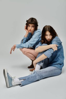 Kot elbiseli iki genç kadın, gri bir arka planda birlikte poz veriyorlar..