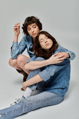 A young lesbian couple in stylish denim attire poses against a grey background. clipart