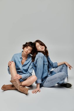 Two young women, clad in denim, pose against a grey backdrop. Lesbian couple clipart