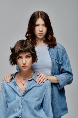 Gri arka planda kot giymiş iki kadın. Bir kadın diğerini kollar, samimiyet ve bağlılık gösterir..