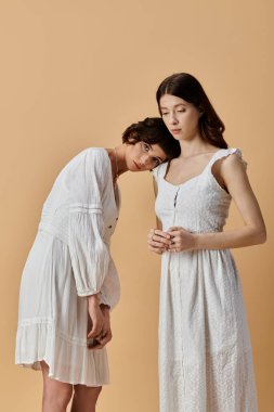 Beyaz yaz elbiseli iki genç kadın bej bir arka planda birbirlerine yakın duruyorlar..