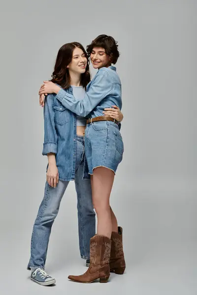 stock image Two young women dressed in denim, embracing and smiling against a grey backdrop.