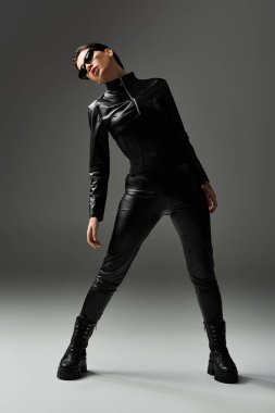 Siyah deri pantolonlu ve çizmeli genç bir kadın gri bir arkaplanda poz veriyor..