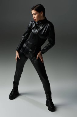 Siyah deri pantolonlu ve ceketli bir kadın gri arka planda poz veriyor..
