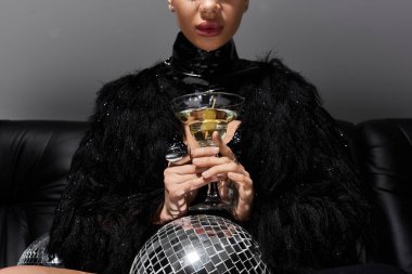 Seksi giyinmiş bir kadın elinde bir martini bardağıyla disko topuyla poz veriyor..