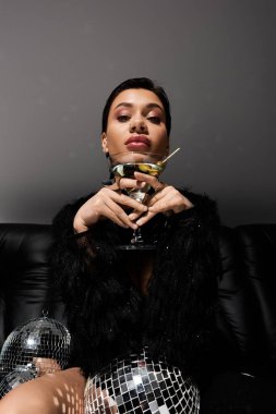 Seksi giyinmiş bir kadın elinde bir martini bardağı ve elinde disko topuyla kanepede oturuyor..