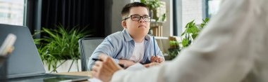 A mother works at her desk in an office, with her son, who has Down syndrome, sitting next to her. clipart