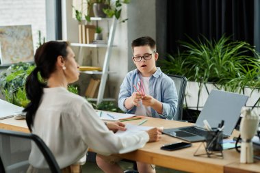 Down sendromlu oğlu yanındaki masada renkli kalemlerle oynarken bir anne dizüstü bilgisayarında çalışıyor..