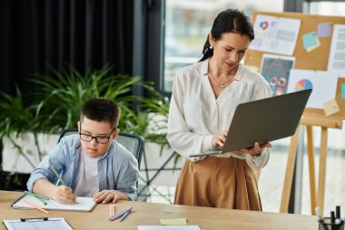 Bir anne dizüstü bilgisayarında çalışırken oğlu Down sendromlu oğlu ofis masasındaki bir defterde renk değiştiriyor..