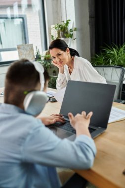 A mother looks on as her son, who has Down syndrome, works on a laptop in their office. clipart