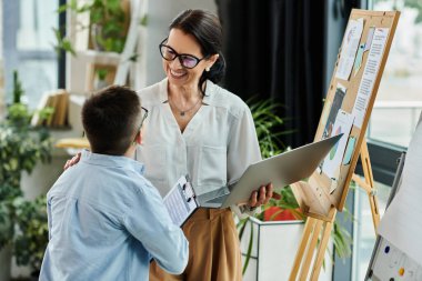 A mother smiles at her son with Down syndrome as they work together in an office setting. clipart