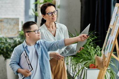 Mother and son in office. Son, with Down syndrome, points to chart on easel while mother watches. clipart