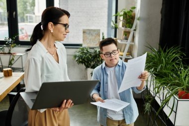 A mother works on her laptop while her son with Down syndrome reviews papers in an office setting. clipart