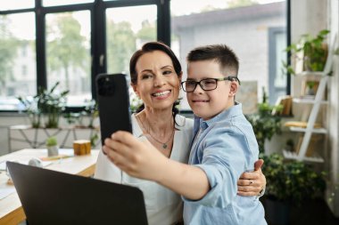 A mother working in her office takes a selfie with her son, who has Down syndrome, demonstrating a positive and inclusive work environment. clipart