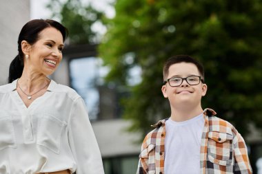 A mother and her son with Down syndrome walk outdoors near an office building, showcasing diversity and inclusion. clipart