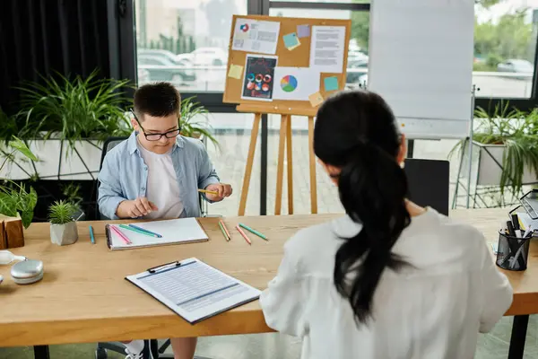 Down sendromlu oğlu yanında çizerken bir anne bir ofiste masa başında çalışıyor ve aile hayatını ve kapsayıcılığını gösteriyor..