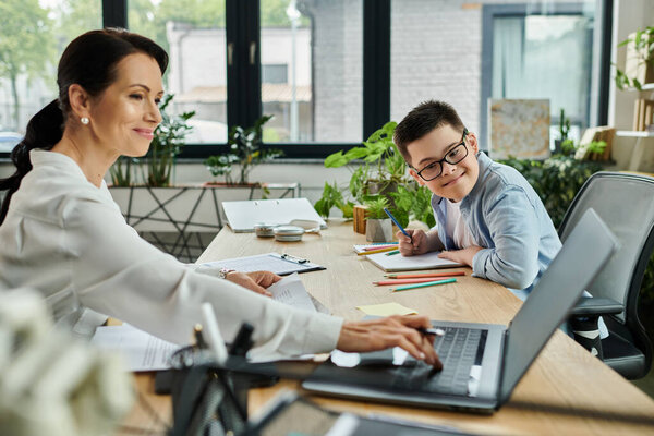 Мать работает на ноутбуке, в то время как ее сын, страдающий синдромом Дауна, рисует за соседним столом.