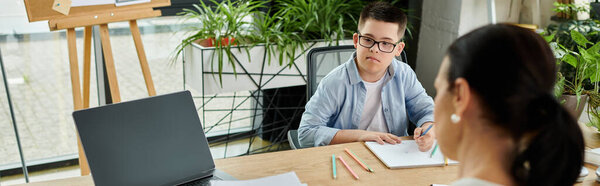 Мать работает над своим ноутбуком в офисе, пока ее сын с синдромом Дауна рисует за столом.