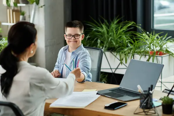 Stock image A mother works on her laptop while her son with Down syndrome sits beside her, drawing with colored pencils.