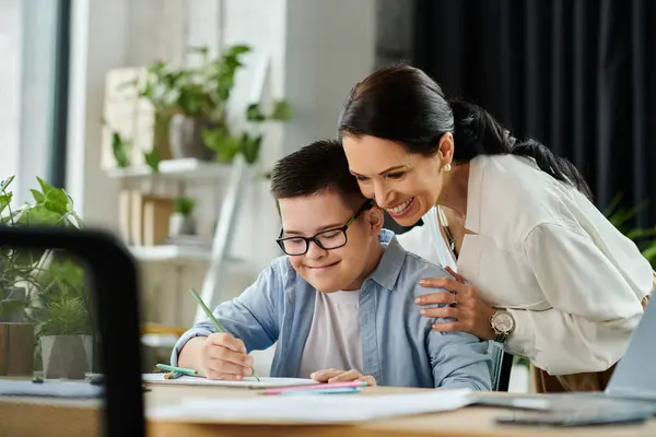 stock image A mother smiles down at her son with Down syndrome as he works on a project at their home office.