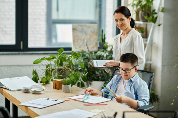 Мать работает над своим ноутбуком, в то время как ее сын, у которого синдром Дауна, рисует за столом в домашнем офисе.