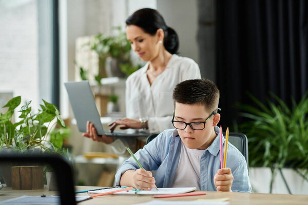 Мать работает на своем ноутбуке, в то время как ее сын с синдромом Дауна окрашивается в блокнот за столом.