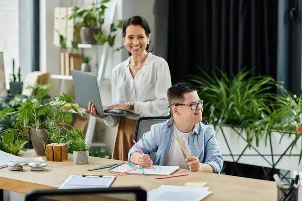 Stock image A mother works on her laptop while her son with Down syndrome draws at a desk in a modern office.