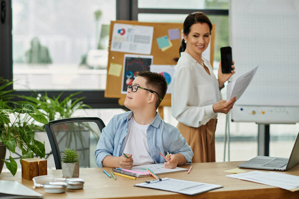 A mother works at her desk in an office while her son with Down Syndrome sits beside her.