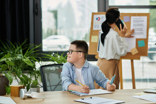 A young boy with Down syndrome sits at a desk in an office setting, working on a project while his mother works nearby.