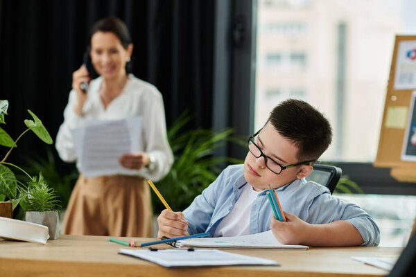A boy with Down syndrome colors at a desk while his mother works in the background.