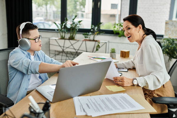 A mother and her son with Down syndrome work side-by-side at a desk in a modern office setting.
