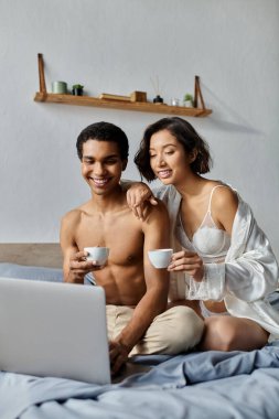 A young interracial couple relaxes in their bedroom, enjoying coffee and looking at a laptop. clipart