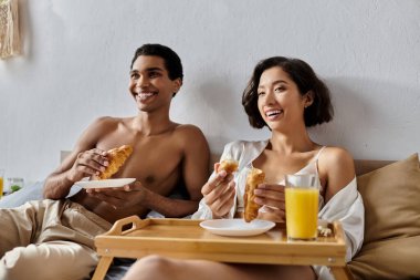 A young multicultural couple enjoys breakfast in bed, laughing and sharing a lighthearted moment together. clipart