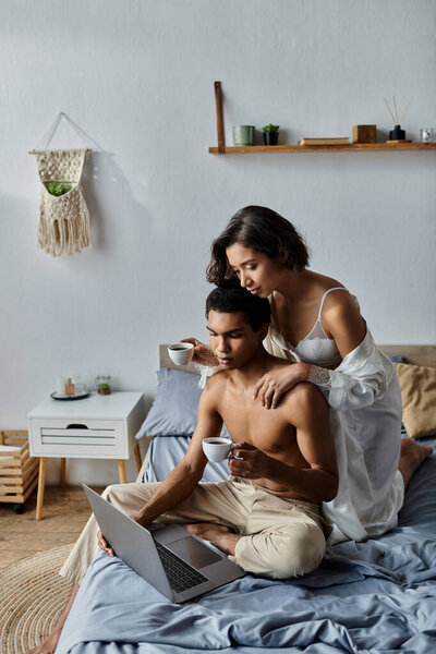 Молодая пара наслаждается уютным утром вместе в своей спальне, потягивая кофе и просматривая на ноутбуке.