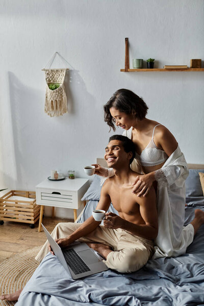 Молодая пара отдыхает в постели, наслаждаясь утренним кофе и работая на ноутбуке.