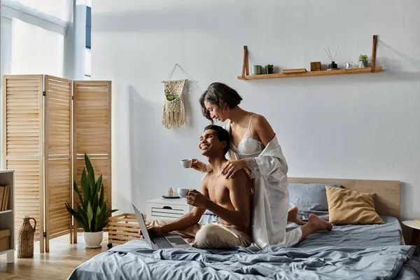 stock image A young multicultural couple enjoys a cozy morning in their bedroom, sharing a cup of coffee and working on a laptop.