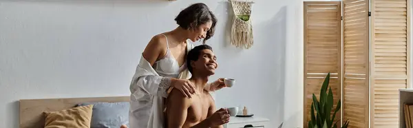 stock image A young multicultural couple spends time together in a bedroom. The woman is sitting on a bed while the man rests his head on her shoulder.