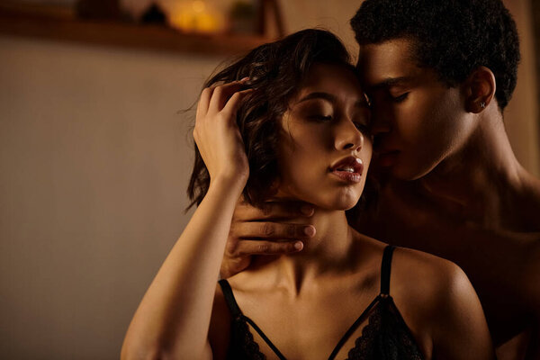 A young Asian woman in black lingerie leans into an African American man on a bed.