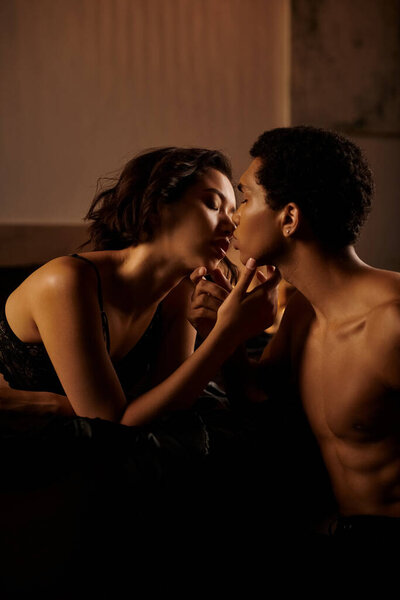 A young Asian woman in black lingerie passionately kisses an African American man on a bed.
