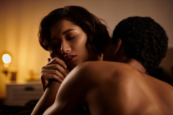 A close-up shot of a young, interracial couple embracing intimately in a dimly lit bedroom, with the womans hand caressing the mans back.