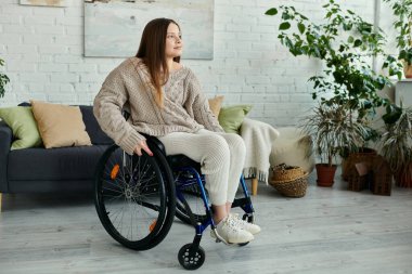 A young woman sits in a wheelchair at home, looking thoughtfully toward the window. clipart