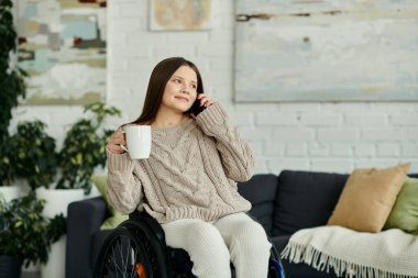Genç bir kadın evinde tekerlekli sandalyesinde oturuyor, bir fincan kahve içip telefon görüşmesi yapıyor..