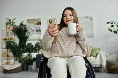Tekerlekli sandalyede oturan genç bir kadın elinde bardak tutarken telefonuna bakıyor..