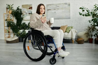 Genç bir kadın evde tekerlekli sandalyesinde oturuyor, telefonunu kullanıyor ve bir fincan kahvenin tadını çıkarıyor..