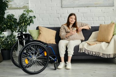 A young woman sits on a sofa in her living room, looking at her phone while her wheelchair rests nearby. clipart