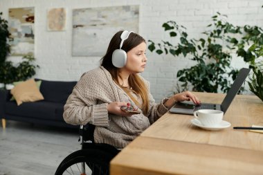 Tekerlekli sandalyedeki genç bir kadın evde dizüstü bilgisayarıyla çalışıyor. Kulaklık takıyor ve elinde telefon var..