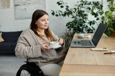 Tekerlekli sandalyedeki genç bir kadın rahat bir evde dizüstü bilgisayarıyla çalışırken bir fincan çay içiyor..